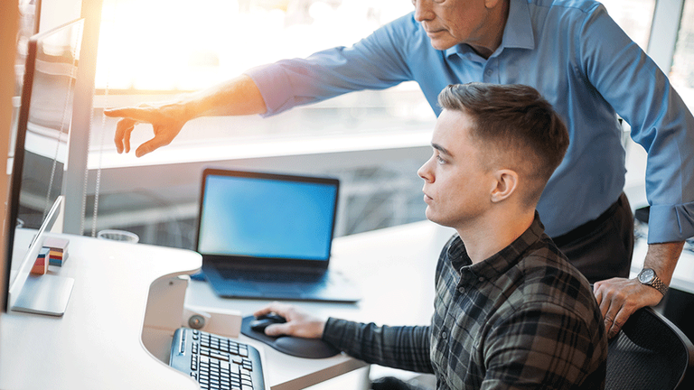 Junger Mann arbeitet am Computer, ein älterer Mann dahinter zeigt ihm etwas auf dem Monitor